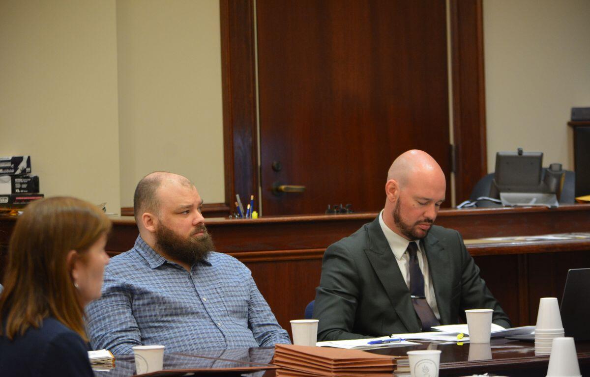 Three people sit at a wood-paneled conference table in a courtroom, with papers and coffee cups in front of them.