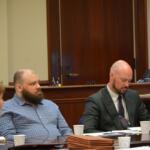 Three people sit at a wood-paneled conference table in a courtroom, with papers and coffee cups in front of them.