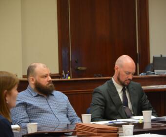 Three people sit at a wood-paneled conference table in a courtroom, with papers and coffee cups in front of them.