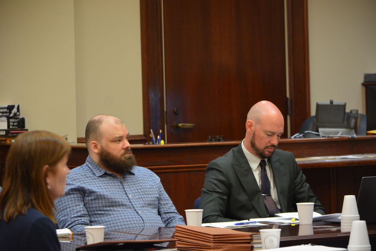 Three people sit at a wood-paneled conference table in a courtroom, with papers and coffee cups in front of them.