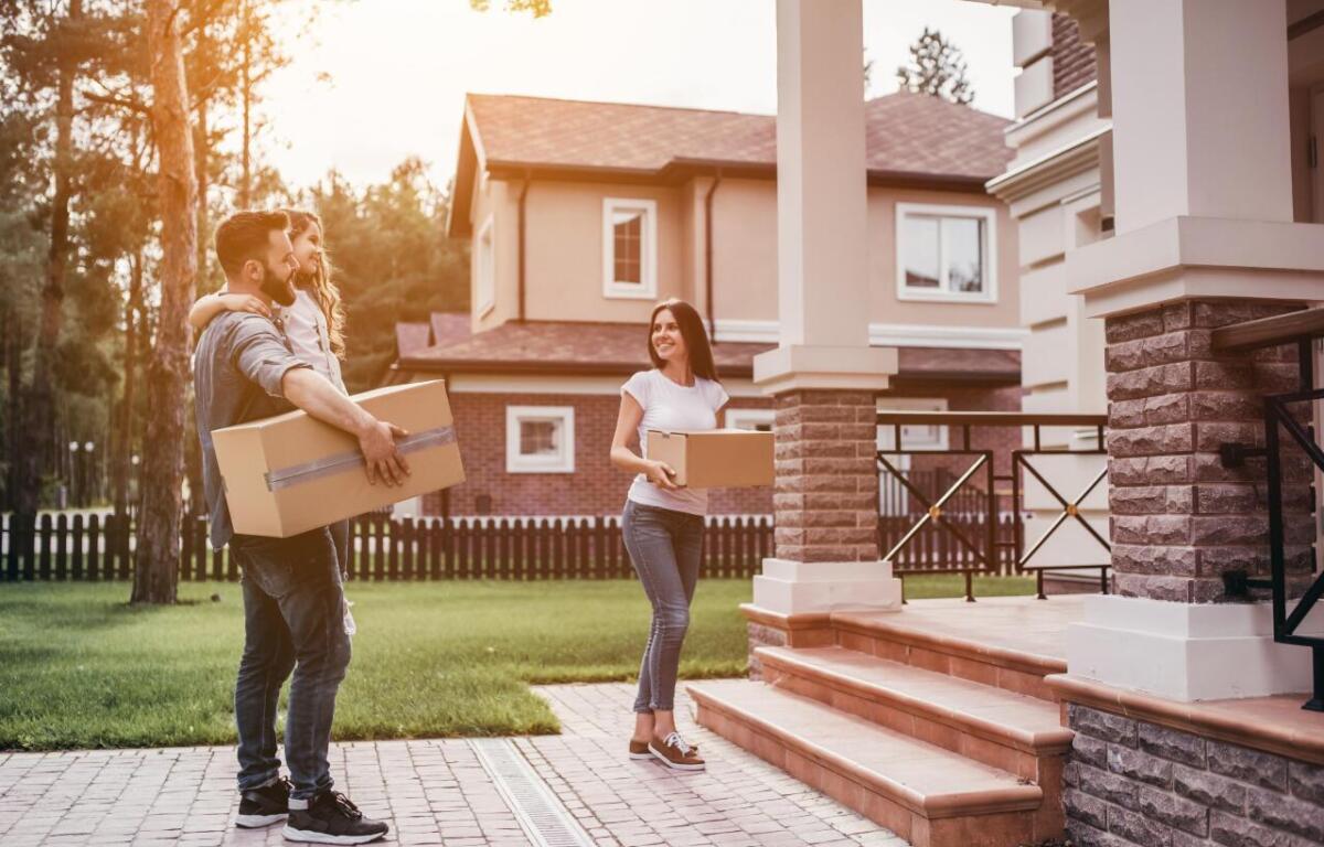 A family carries cardboard boxes toward a large house with a brick-and-stone porch, ready to move in.