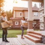 A family carries cardboard boxes toward a large house with a brick-and-stone porch, ready to move in.