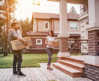 A family carries cardboard boxes toward a large house with a brick-and-stone porch, ready to move in.