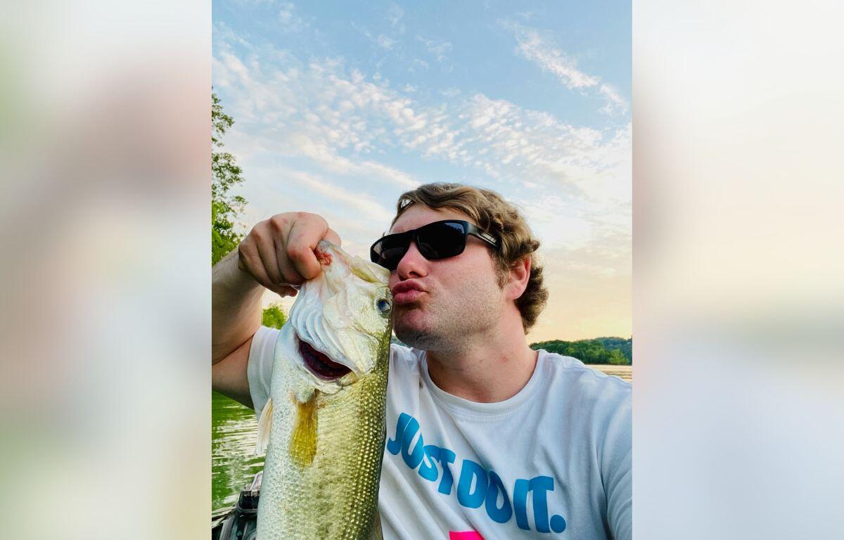 Man in sunglasses kissing a large bass he just caught by a lake at sunset