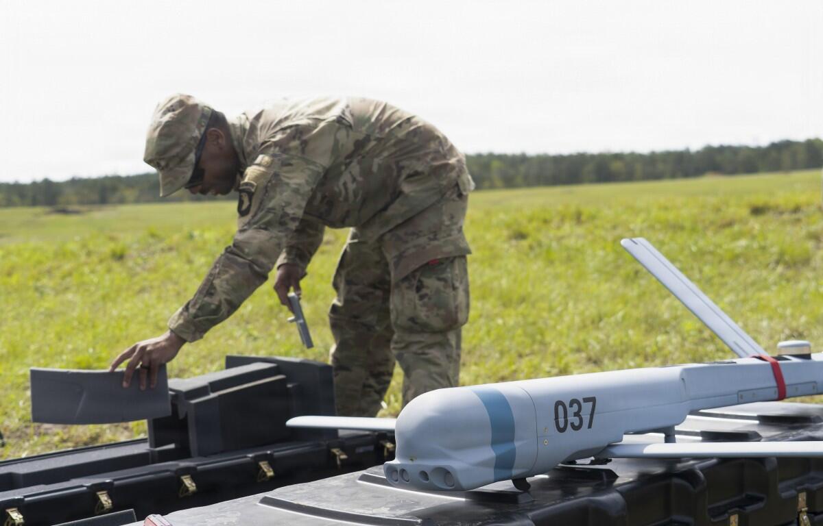 Soldier in camouflage bends over a gray unmanned drone on a field, inspecting its fuselage.