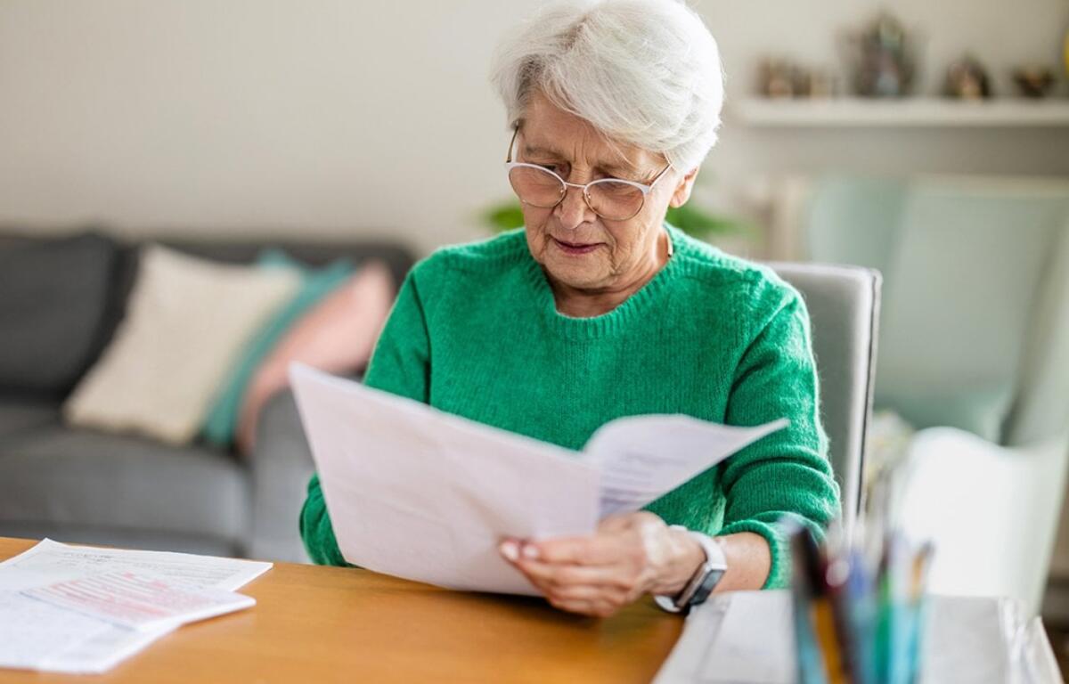 Elderly woman with short white hair and glasses reads papers at a wooden table in a bright living room, wearing a green sweater.