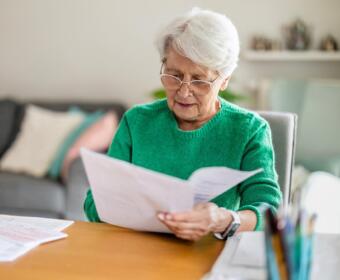 Elderly woman with short white hair and glasses reads papers at a wooden table in a bright living room, wearing a green sweater.
