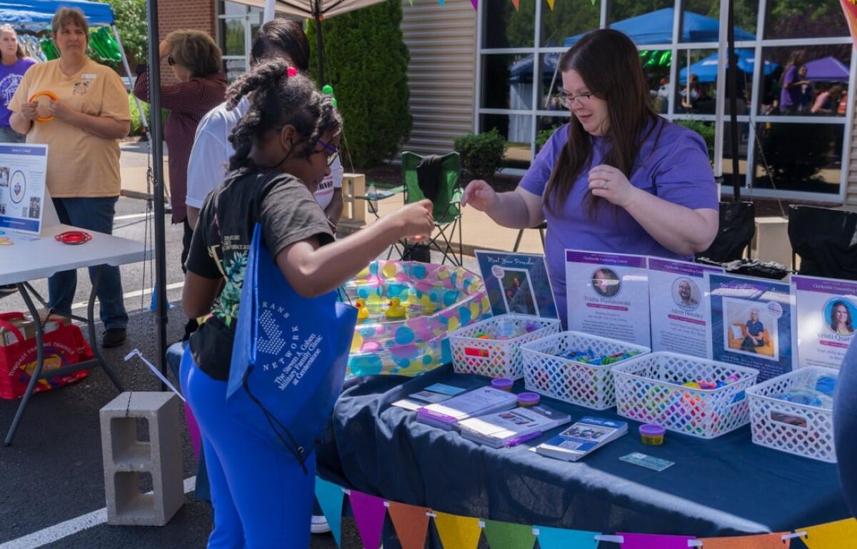 A woman in a purple shirt hands an item to a young girl at a booth with colorful banners and baskets of supplies outside a building.