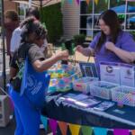 A woman in a purple shirt hands an item to a young girl at a booth with colorful banners and baskets of supplies outside a building.