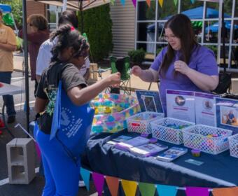 A woman in a purple shirt hands an item to a young girl at a booth with colorful banners and baskets of supplies outside a building.