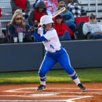 Softball batter in a white and blue uniform swings at the plate with a helmet on, spectators behind the fence.
