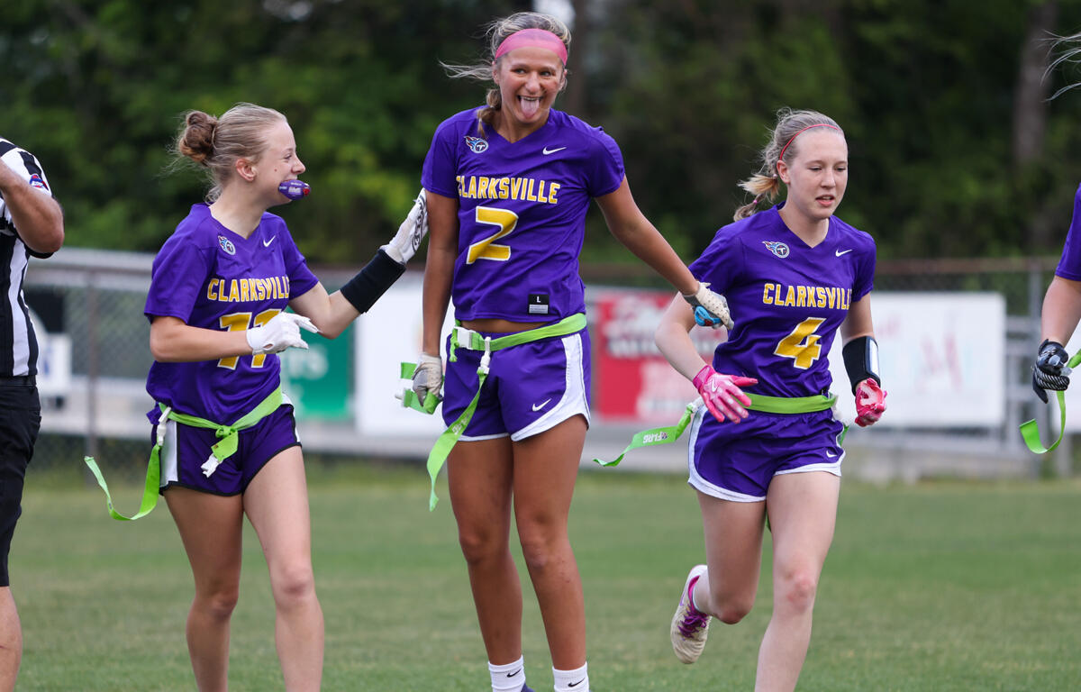 Three Clarksville lacrosse players in purple uniforms celebrate on a grassy field, arms linked and smiling.