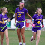 Three Clarksville lacrosse players in purple uniforms celebrate on a grassy field, arms linked and smiling.