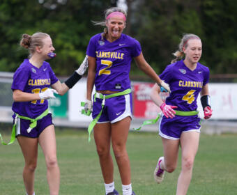 Three Clarksville lacrosse players in purple uniforms celebrate on a grassy field, arms linked and smiling.