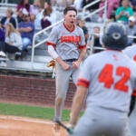 Baseball player in a Cobra gray uniform celebrates on the dirt field with clenched fists while teammates approach and fans watch from the stands.