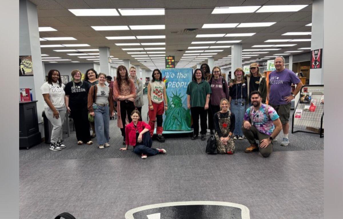 Group of about 16 people posing for a photo in a library, centered around a blue banner that says 'We the People' with a Statue of Liberty image.