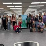 Group of about 16 people posing for a photo in a library, centered around a blue banner that says 'We the People' with a Statue of Liberty image.