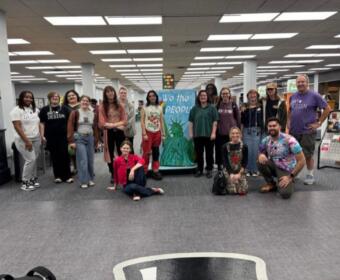 Group of about 16 people posing for a photo in a library, centered around a blue banner that says 'We the People' with a Statue of Liberty image.