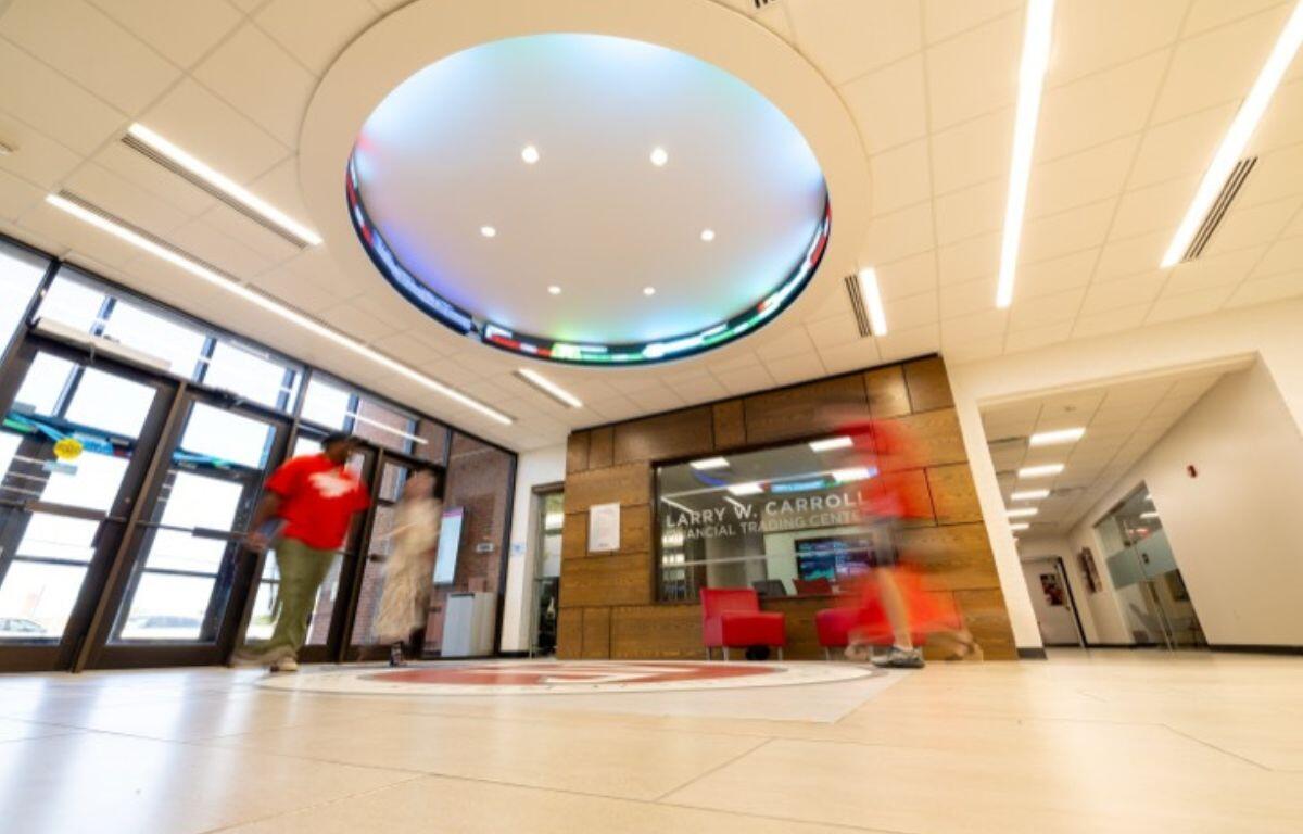 Interior lobby of a modern building with a circular illuminated ceiling light and glass double doors at left; reception area with a wooden wall and sign reading 'LARRY W. CARROLL FINANCIAL TRADING CENTER' in the background.