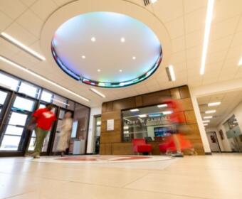 Interior lobby of a modern building with a circular illuminated ceiling light and glass double doors at left; reception area with a wooden wall and sign reading 'LARRY W. CARROLL FINANCIAL TRADING CENTER' in the background.