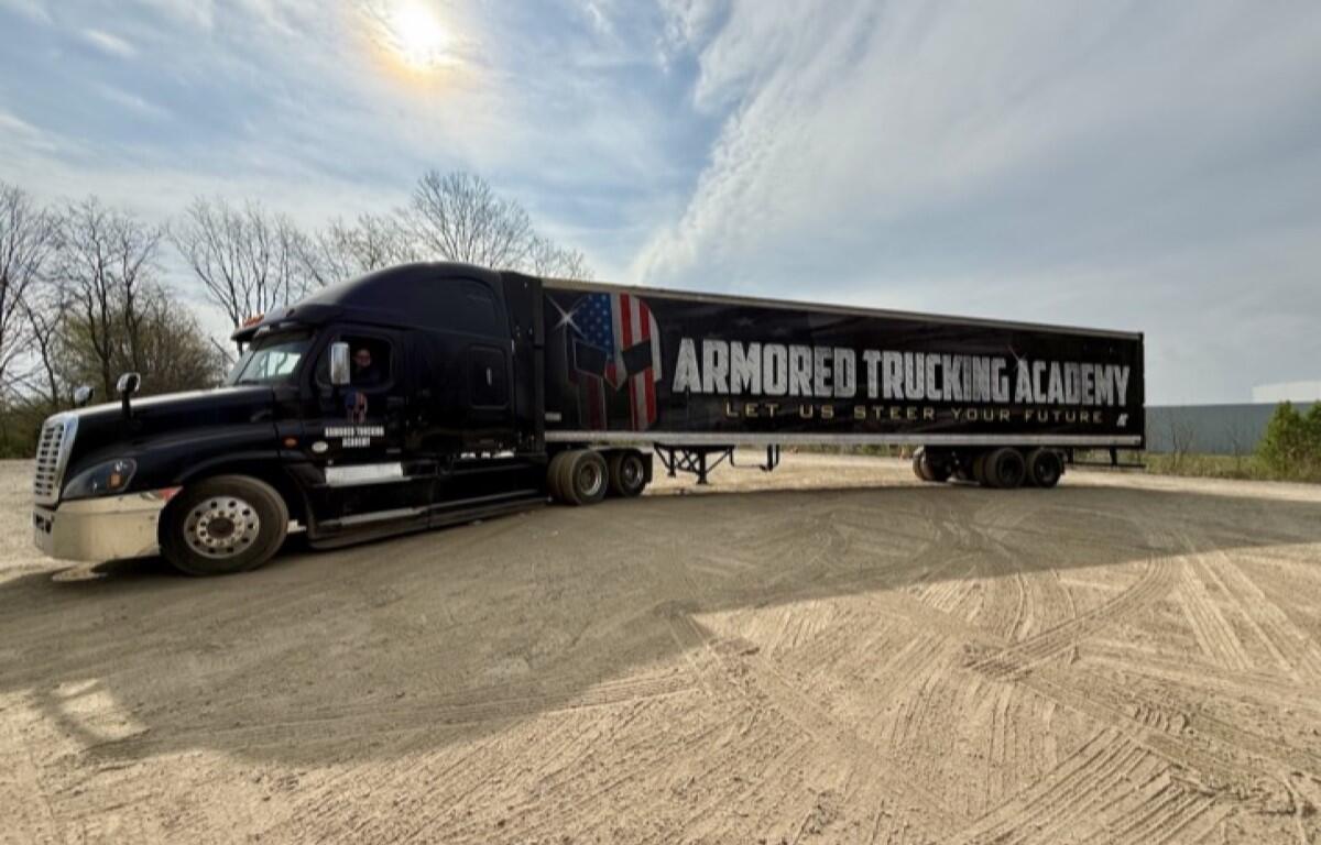 Black semi-truck with a long trailer reading 'Armored Trucking Academy' parked on a dirt lot under a cloudy sky, with the slogan 'Let us steer your future' visible on the trailer.