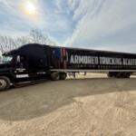 Black semi-truck with a long trailer reading 'Armored Trucking Academy' parked on a dirt lot under a cloudy sky, with the slogan 'Let us steer your future' visible on the trailer.