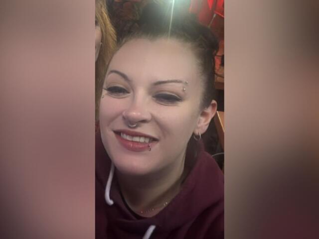 Smiling woman with a nose ring and eyebrow piercing wearing a maroon hoodie.
