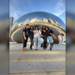 Five friends jump in unison in front of Chicago's Cloud Gate sculpture (The Bean), with the city skyline reflected on its curved surface.
