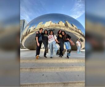 Five friends jump in unison in front of Chicago's Cloud Gate sculpture (The Bean), with the city skyline reflected on its curved surface.