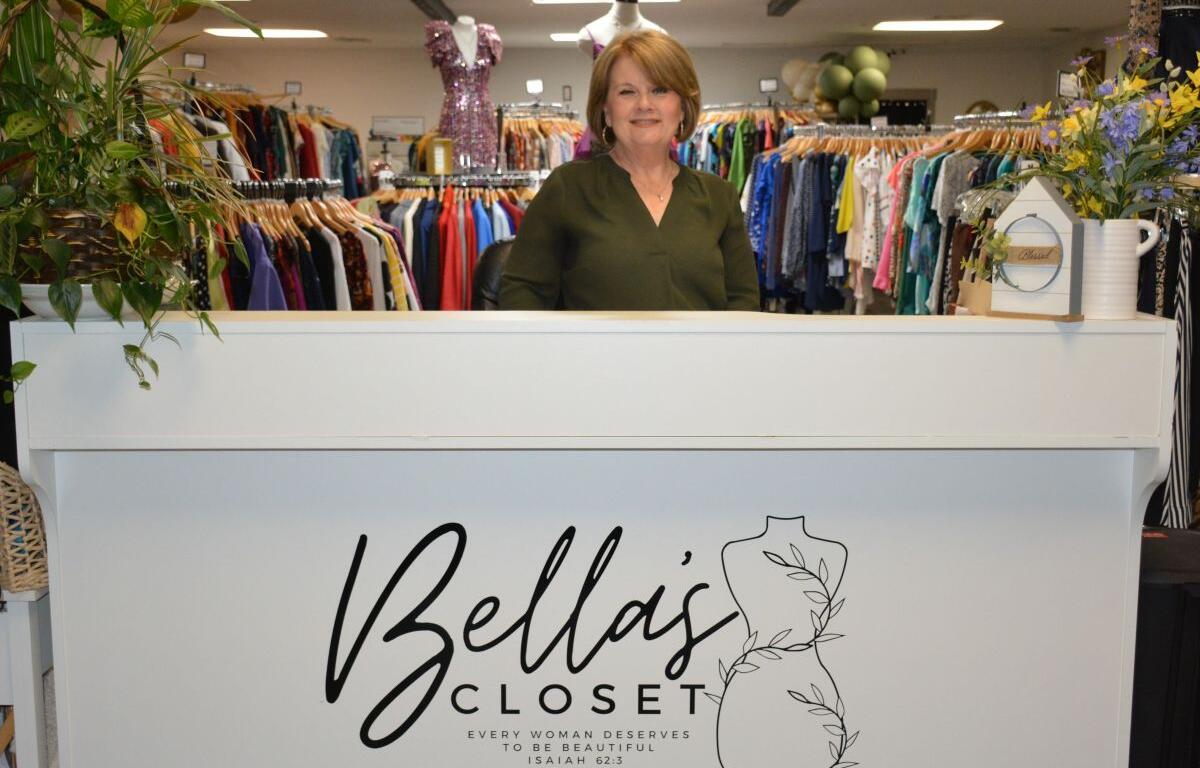 Smiling woman stands behind a white front desk at Bella's Closet, a women’s clothing store.