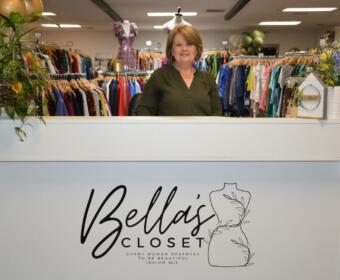 Smiling woman stands behind a white front desk at Bella's Closet, a women’s clothing store.