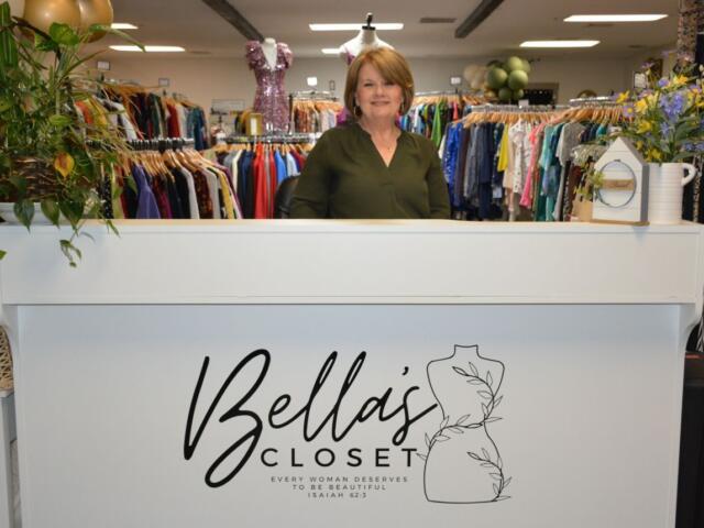 Smiling woman stands behind a white front desk at Bella's Closet, a women’s clothing store.