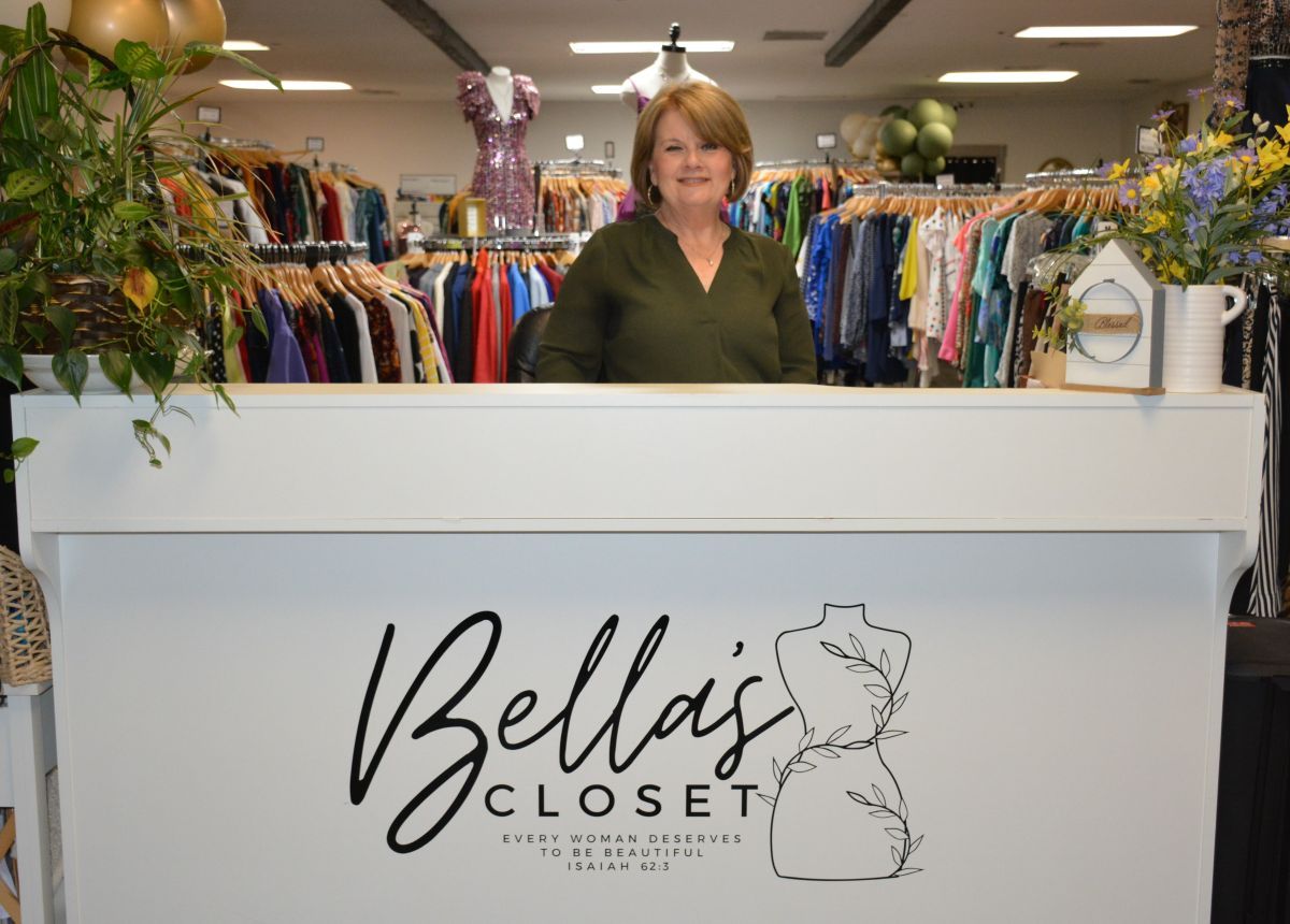 Smiling woman stands behind a white front desk at Bella's Closet, a women’s clothing store.