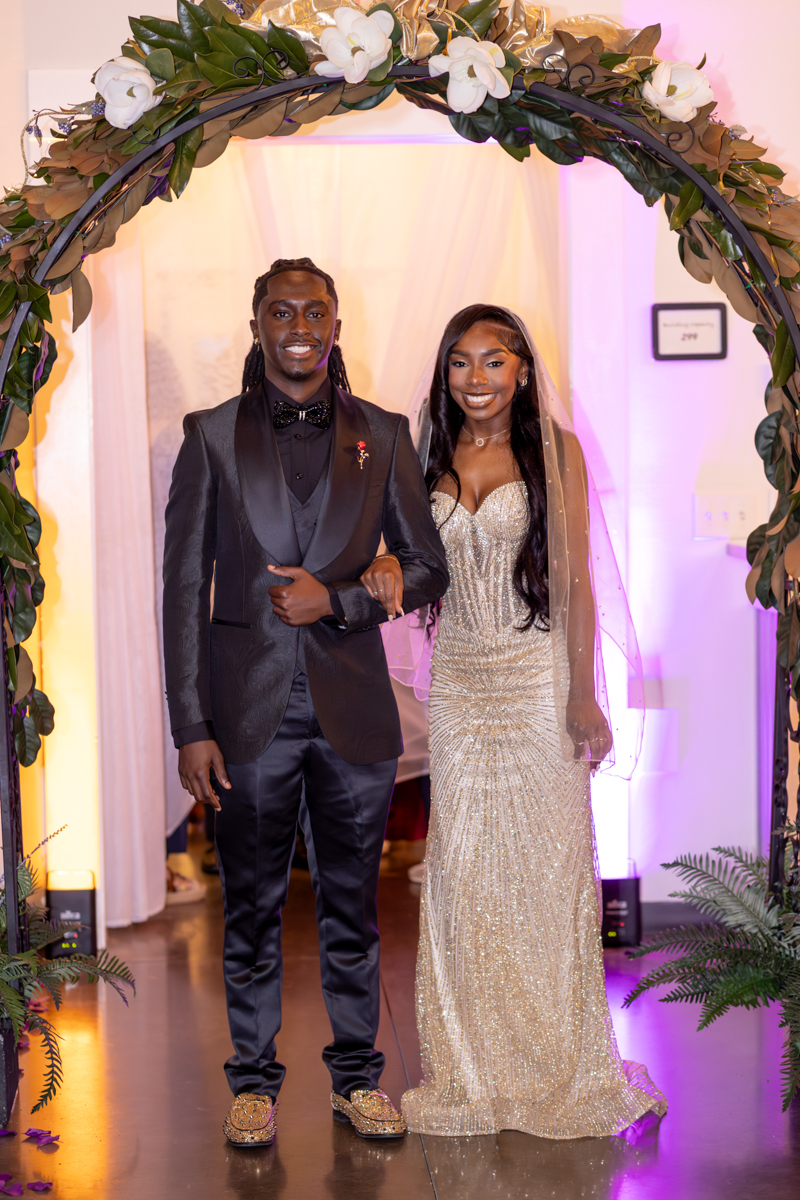 Couple walking arm in arm under a floral arch at a wedding; man in black suit and bow tie, woman in a sparkling champagne gown.