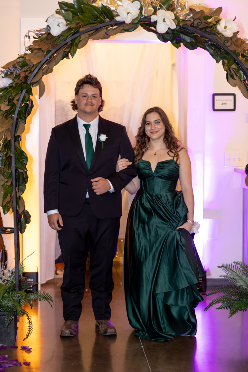 Couple in formal wear under a flower arch: man in black suit with green tie, woman in emerald gown.