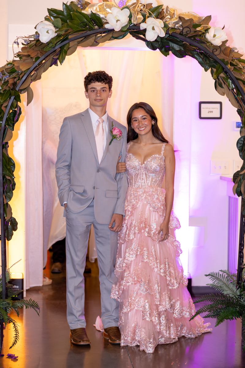 Couple in formal wear pose under a floral arch; she wears a pink lace gown, he a light gray suit, with pink boutonniere, pink-lit venue in the background.