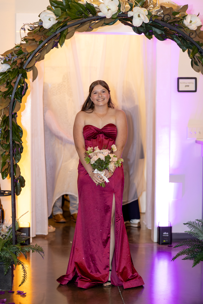 Young woman in a burgundy strapless evening gown holding a bouquet, standing under a floral arch at a formal event