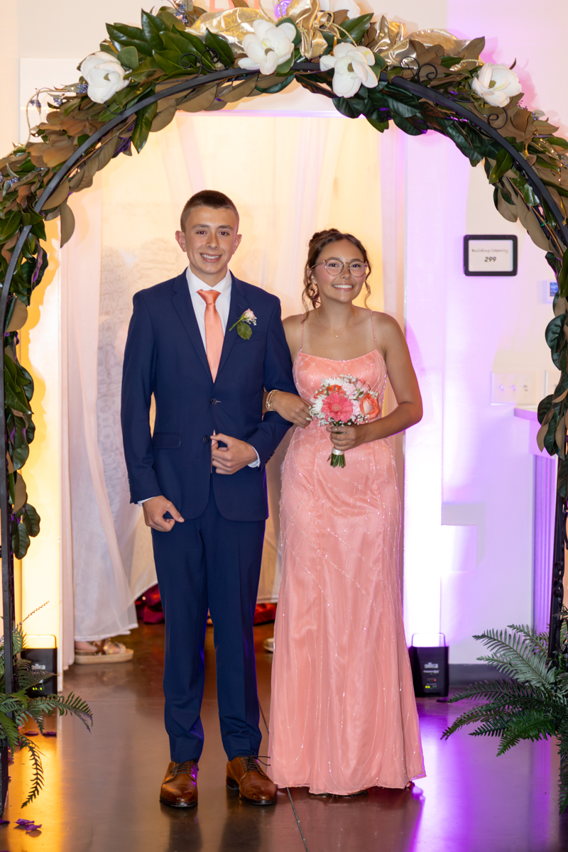 Young couple poses under a flower arch at a formal event; man in a navy suit with peach tie, woman in a pink gown holding a bouquet.