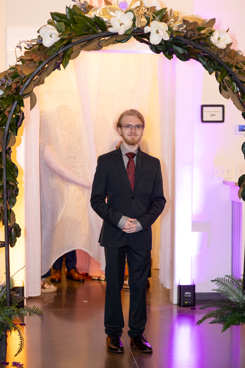 Man in a dark suit and red tie stands with hands clasped under a floral arch at a formal event.