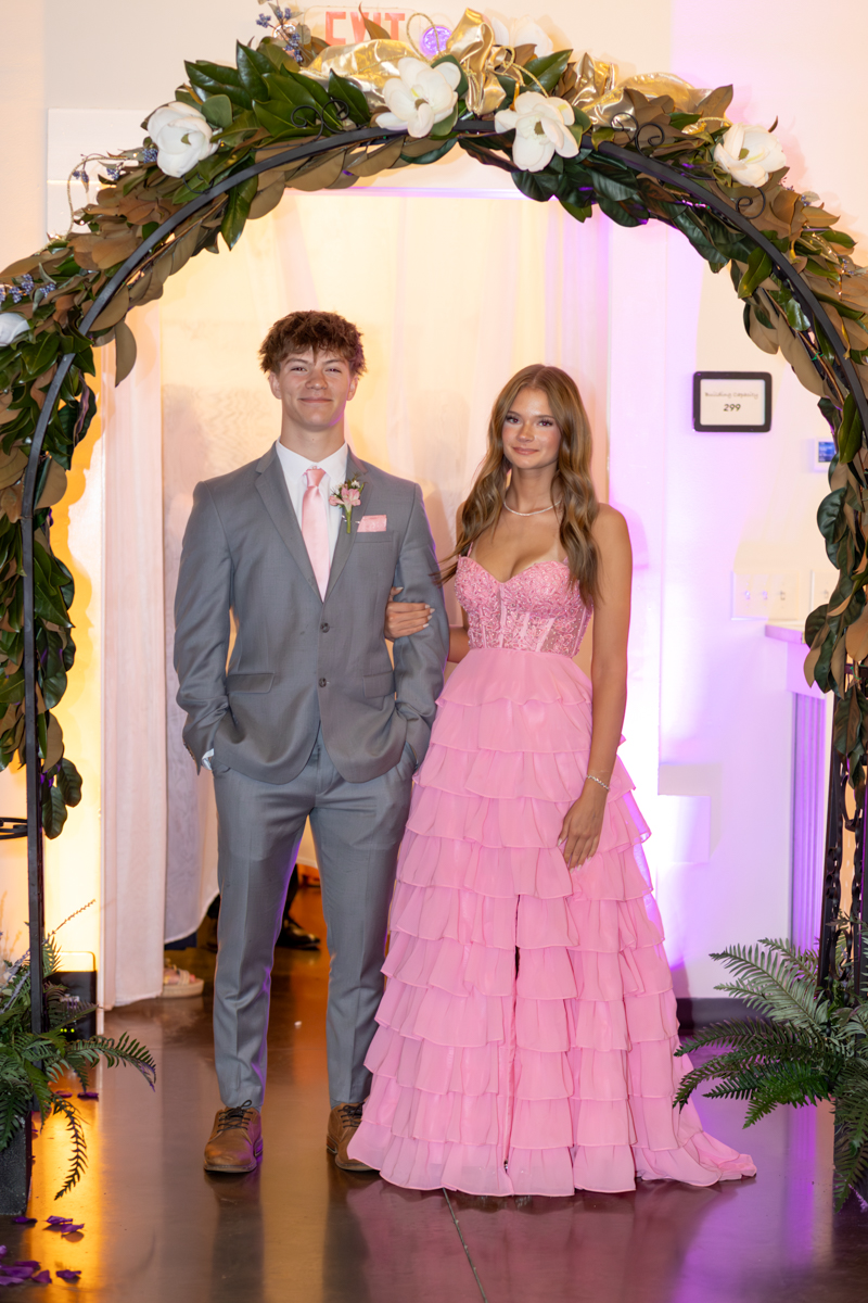Young couple in formal attire posing under a flower arch at a prom-like event.
