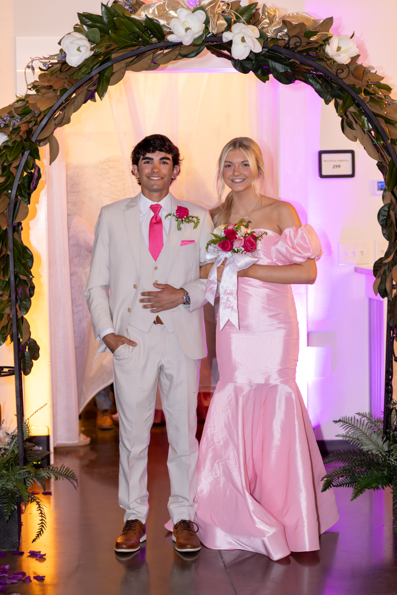 Couple in formal attire posing under a floral arch, pink accents, smiling at the camera.