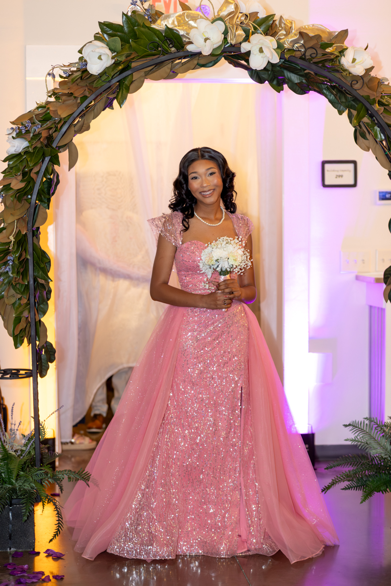 Smiling woman in a pink sequined gown holding a small bouquet, standing under a floral arch at a formal event.