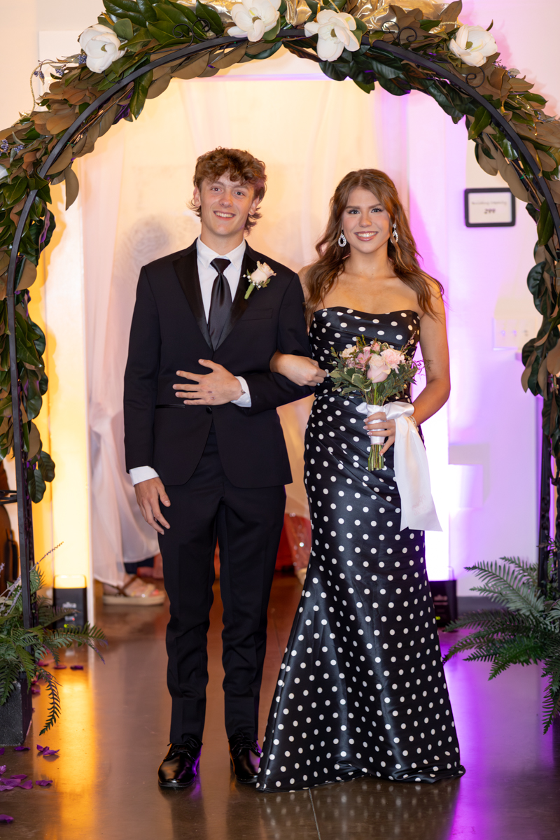 Smiling couple in formal attire posing under a flower arch; man in a black suit, woman in a black polka-dot gown holding a bouquet.