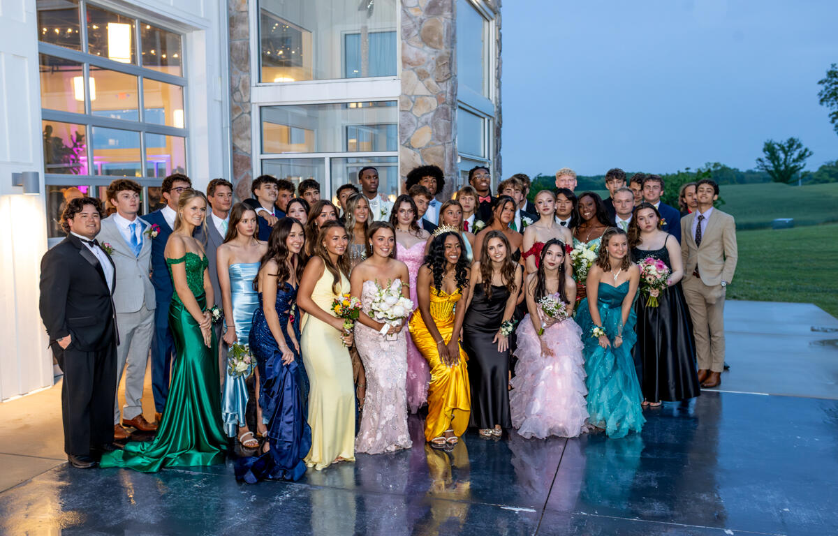 Group of students in formal wear posing together outside a modern glass-and-stone building, likely at prom or formal event at dusk/night.