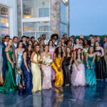 Group of students in formal wear posing together outside a modern glass-and-stone building, likely at prom or formal event at dusk/night.