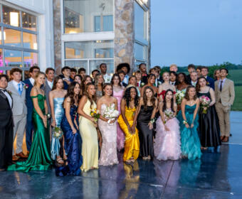 Group of students in formal wear posing together outside a modern glass-and-stone building, likely at prom or formal event at dusk/night.