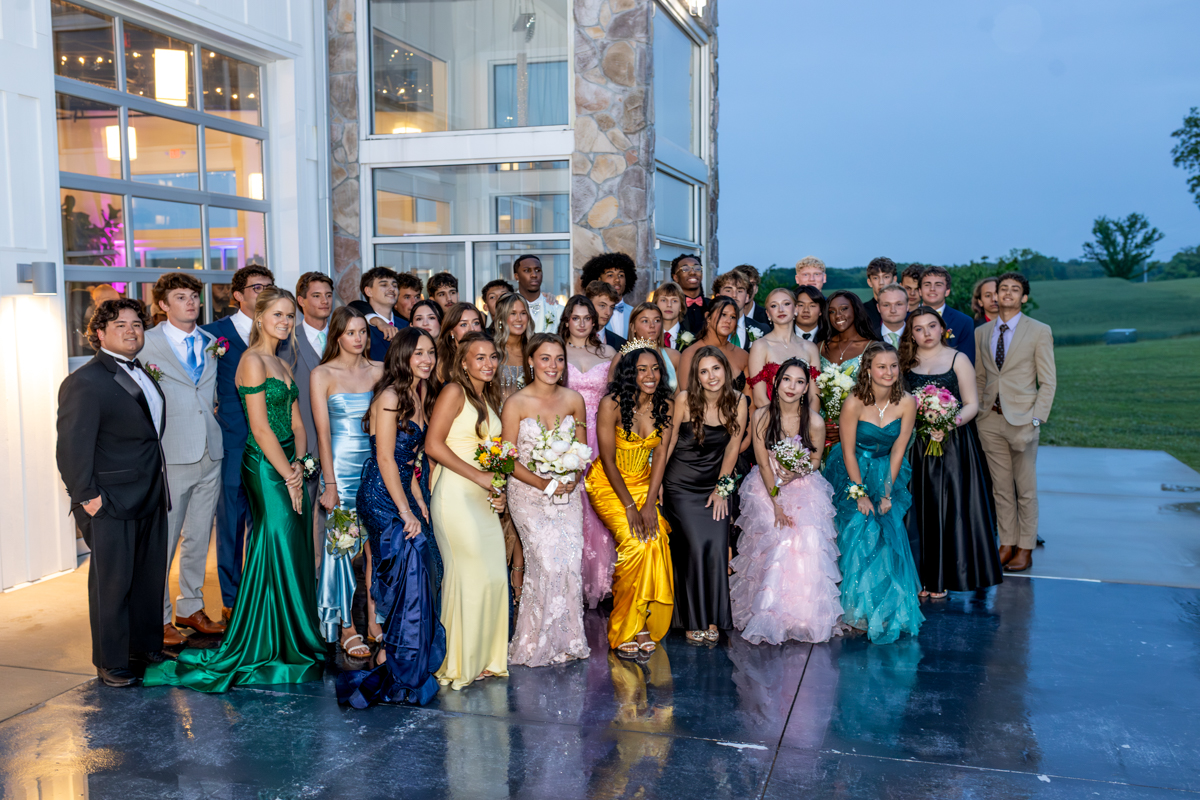 Group of students in formal wear posing together outside a modern glass-and-stone building, likely at prom or formal event at dusk/night.