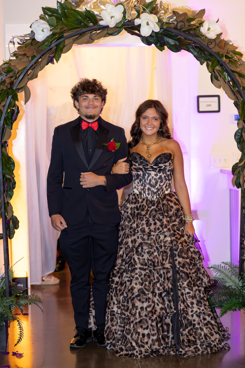 Couple in formal wear posing arm-in-arm under a flower-covered arch at a gala or prom event.