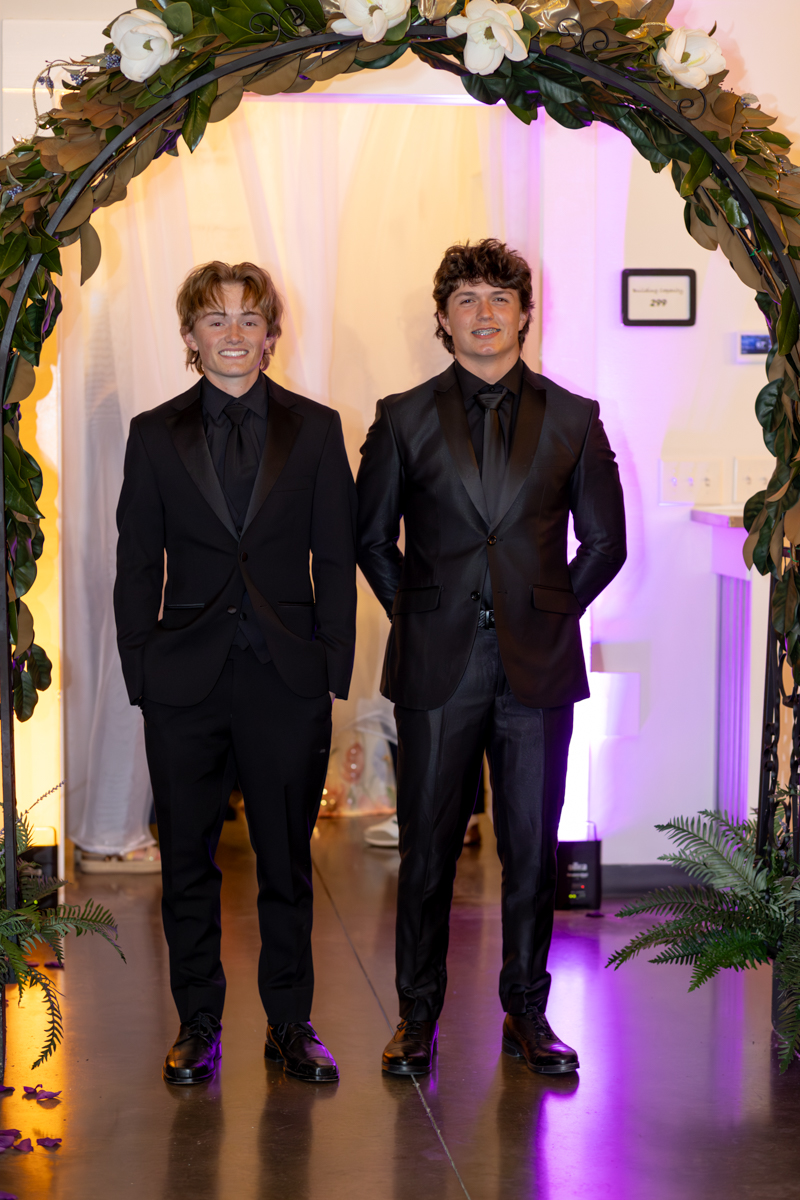 Two men in black tuxedos pose under a flower-decorated arch at an indoor formal event, smiling for the camera.