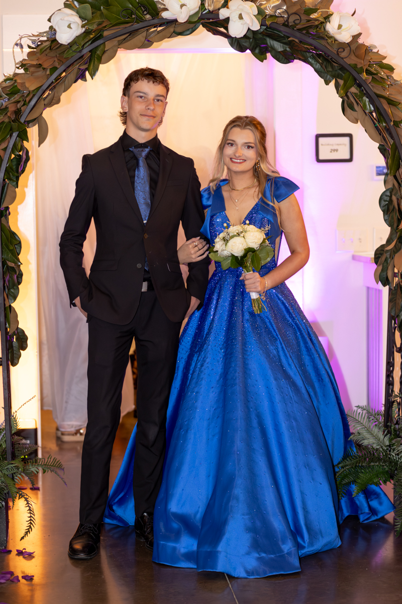 Couple in formal wear posing under a flower arch; man in a black suit and blue tie, woman in a sparkling blue gown holding a bouquet of white roses.